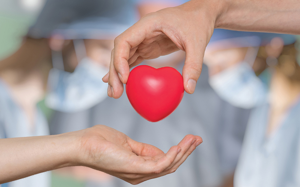 A hand gently passes a red heart to another in front of surgeons in masks and gowns.