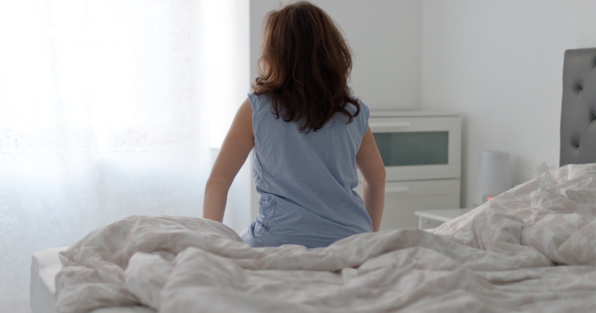 Woman with dark hair sitting on a bed, facing away from the camera.