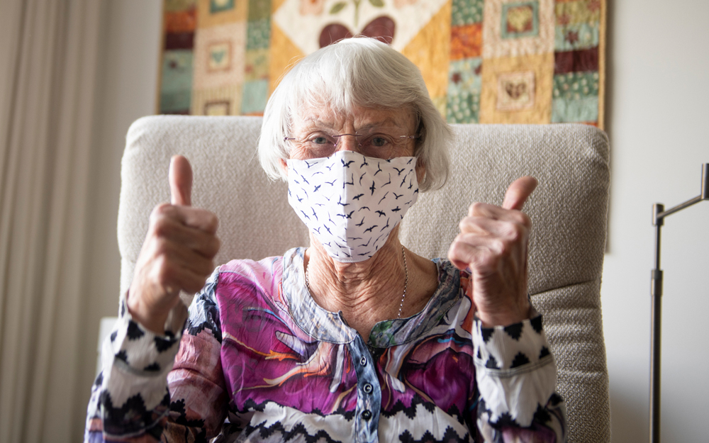 An older adult woman with a mask on to avoid COVID-19 giving the thumbs up after getting help from friends and family.