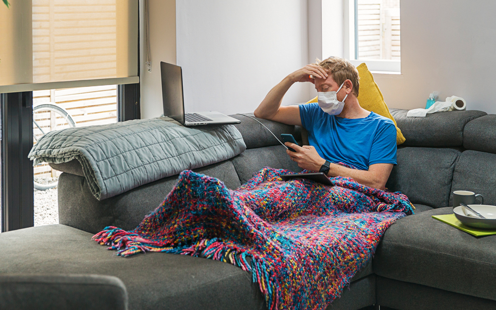 A man sick at home sitting on the couch wondering if he has COVID-19, the flu or a cold.