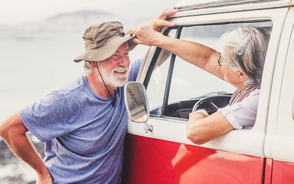 An older man living with epilepsy talks to his wife through the car door while parked at the beach.