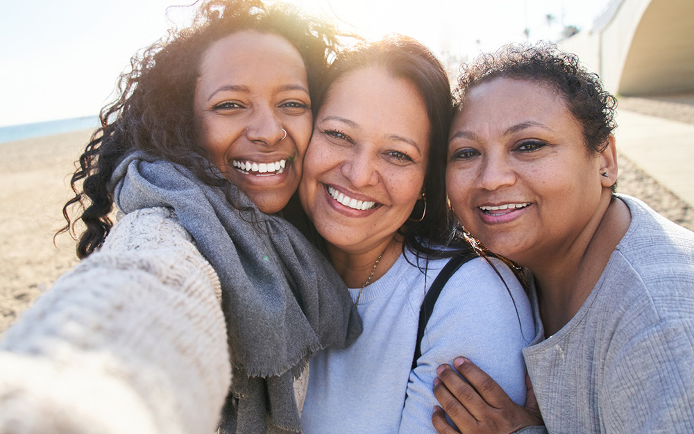 A smiley photo of a family of three women taking a selfie of themselves, looking at camera and with the beach behind them.