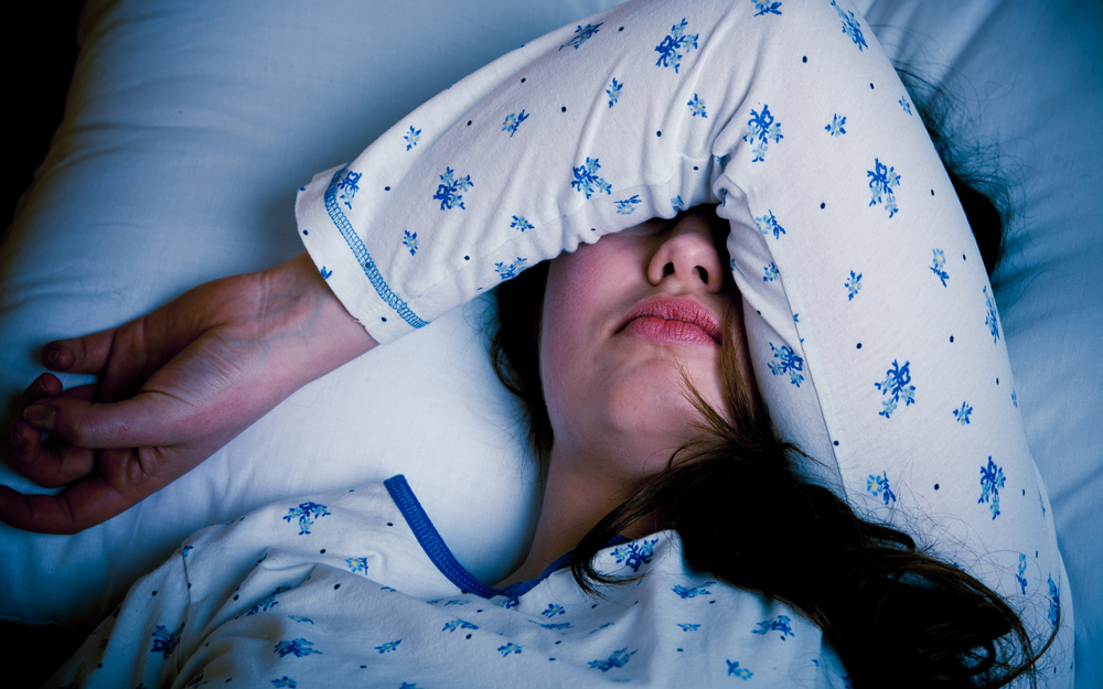 A young woman with a headache, lying in bed with her arm draped over her eyes.