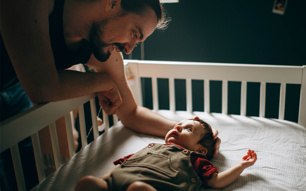 Father interacting with his infant son laying in a crib.