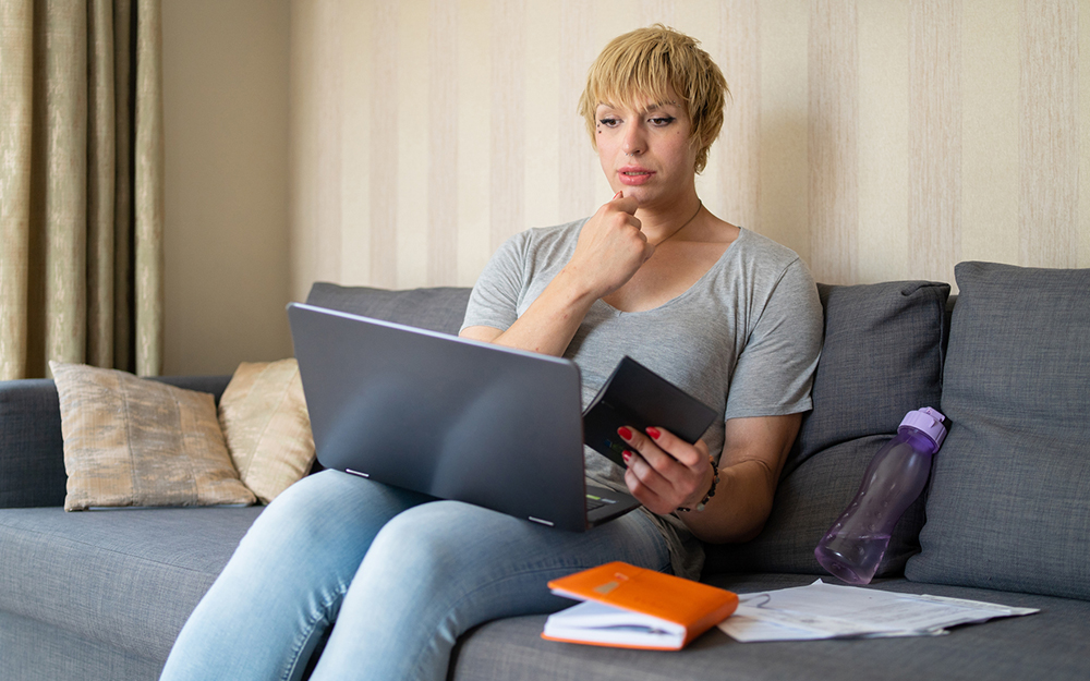 A transitioning woman looking at her laptop while working on her finances sitting on a sofa at home with her paperwork next to her