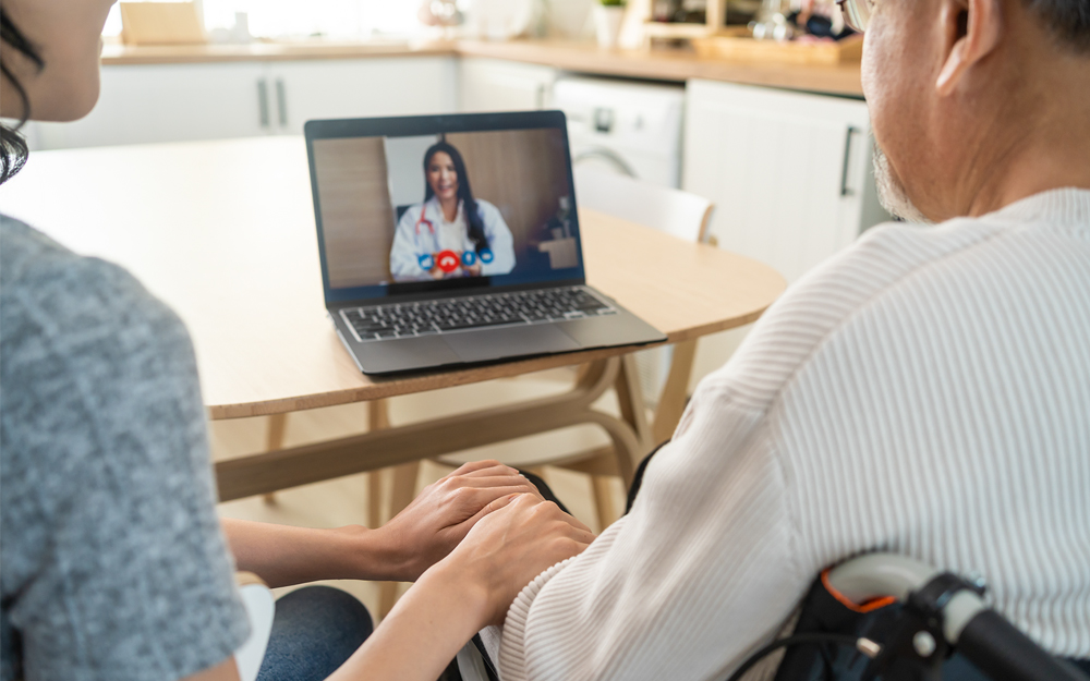 An infirm patient sitting with a care giver while speaking with a physician on a laptop.
