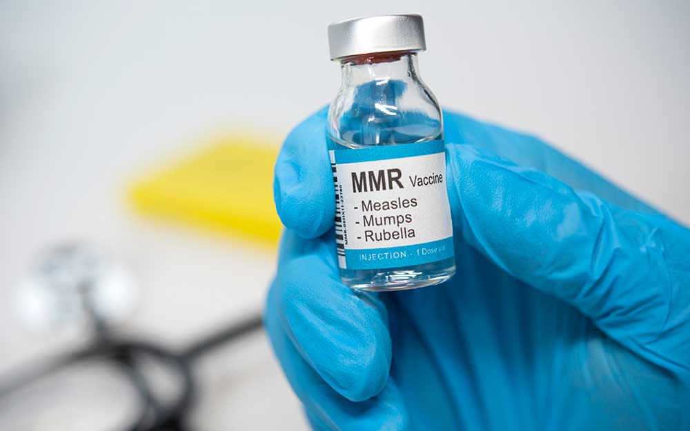 A hand with nitrile gloves and a vial of MMR vaccine.