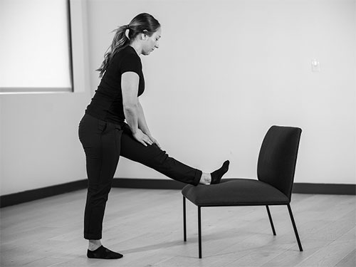 Woman places one heel on a chair, straightening both legs and stand tall.