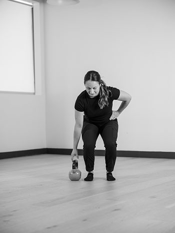 Woman standing with one arm on her side, bending to hold a kettle bell.
