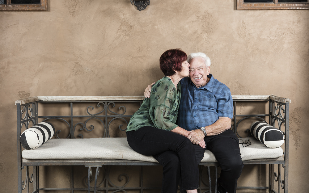 Ruth and Bill Reid sitting together on a couch while Ruth kisses her husband Bill on the cheek after being diagnosed free of throat cancer from HPV.